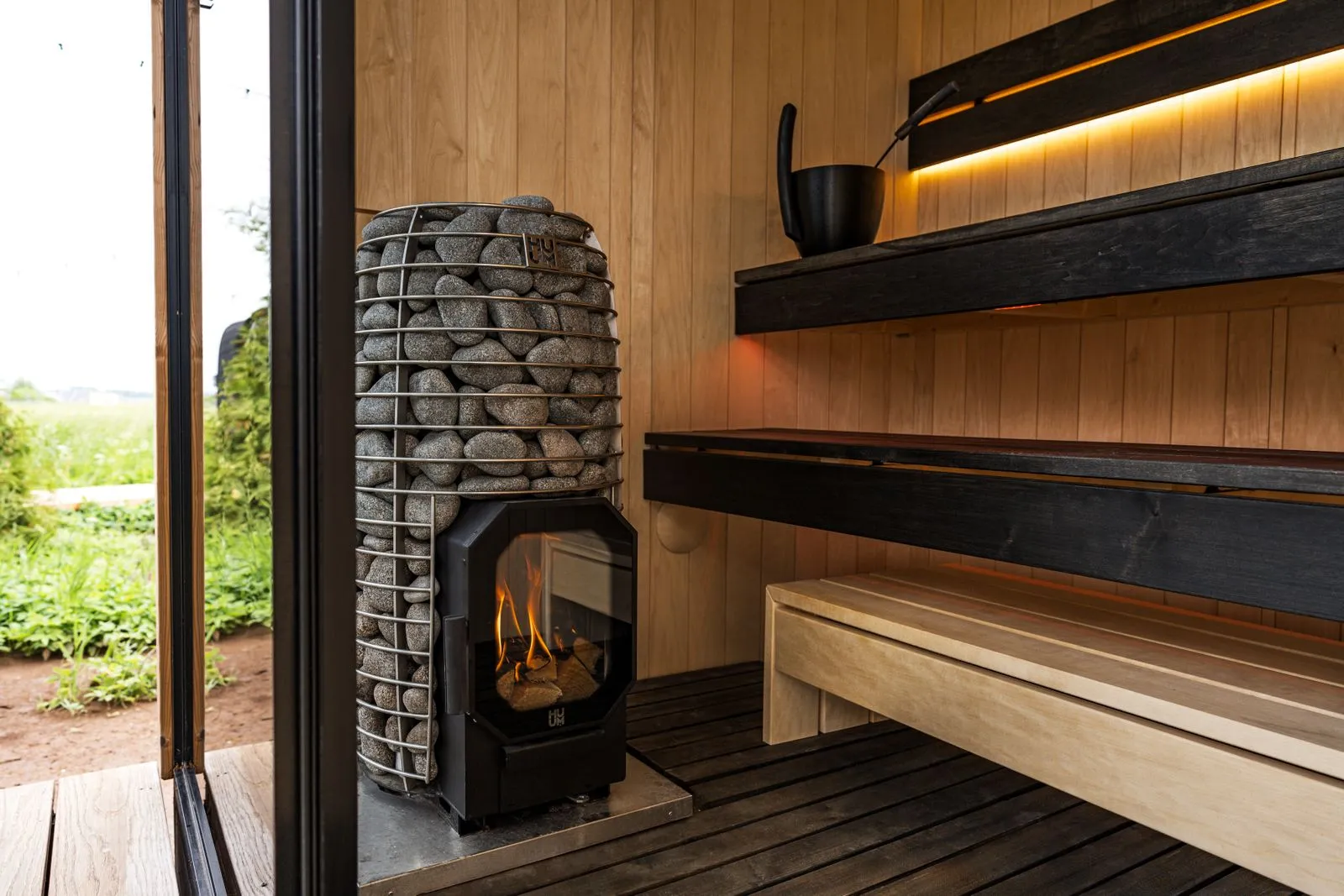 Stone-clad sauna heater with a visible flame beside a wooden bench in a Finnish sauna